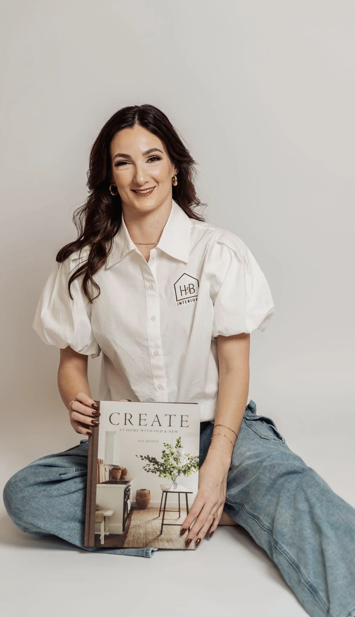 A woman with dark wavy hair, wearing a white shirt with puffed sleeves and jeans, sitting cross-legged while holding a book titled 'Create at Home with Old & New' in front of a plain white background.