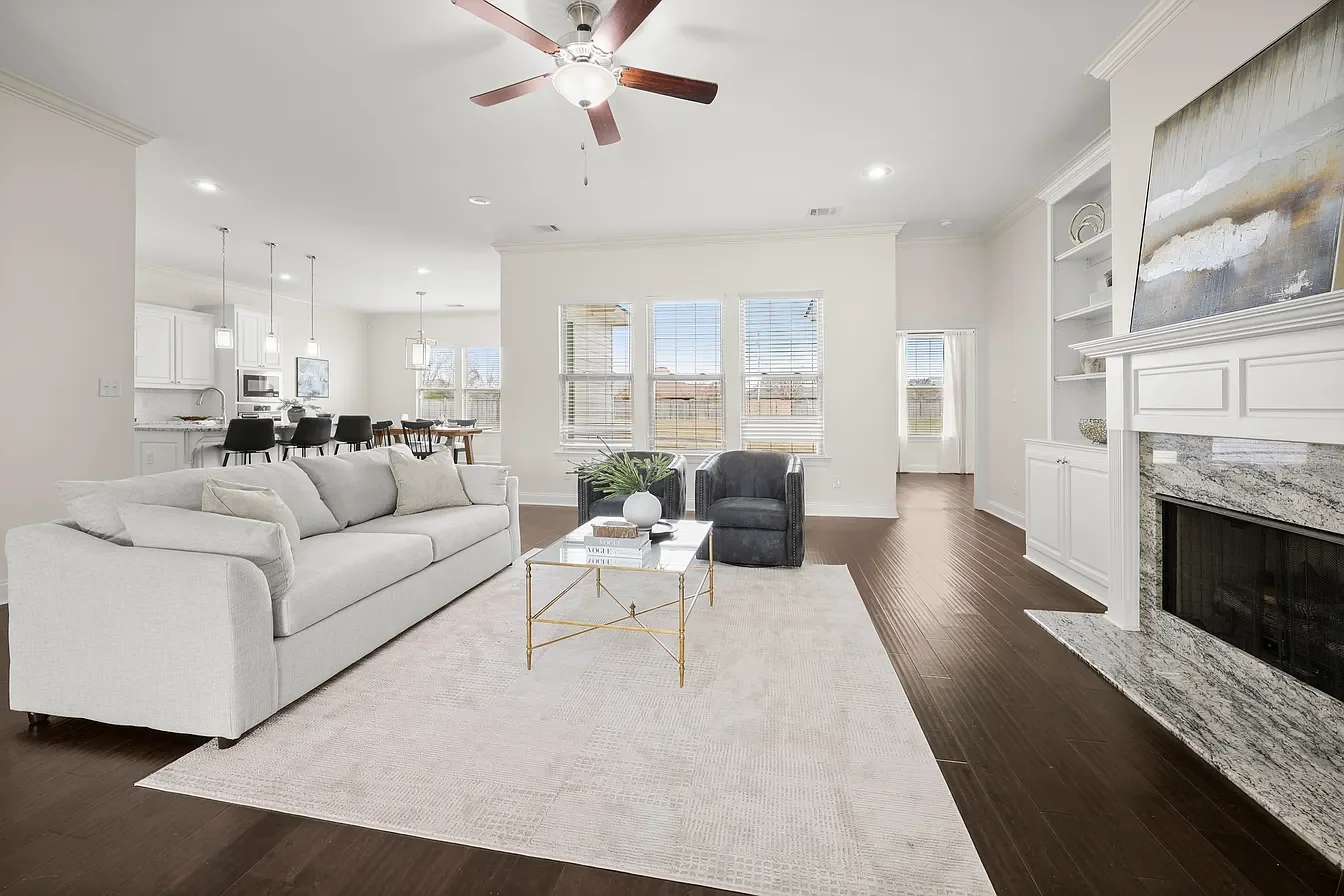 Bright living room with white walls, dark hardwood floors, a white sofa, and a fireplace with a marble mantel. There are two black armchairs, a coffee table with books and a plant, and large windows with white blinds.