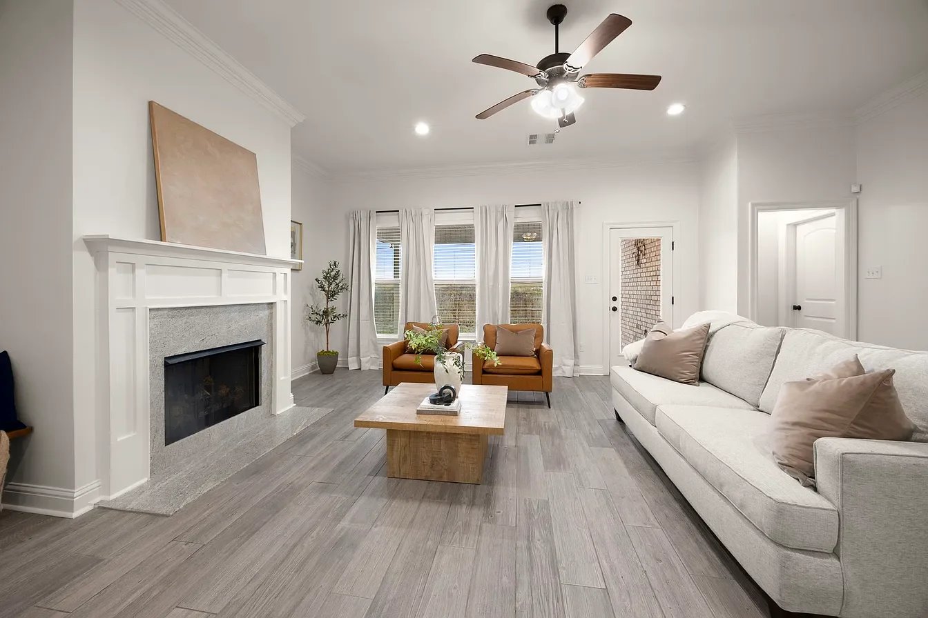 A modern living room with white walls, a white fireplace with a beige painting above, a light gray sofa with beige pillows, a wooden coffee table with decorative items, two tan armchairs, a ceiling fan, and large windows with white curtains.