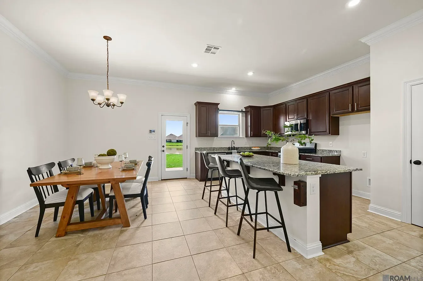Kitchen with dark wooden cabinets, granite countertops, a small island with bar stools, a dining table set with chairs, windows showing outdoor view, tiled floor, and a vase with greenery on the island.