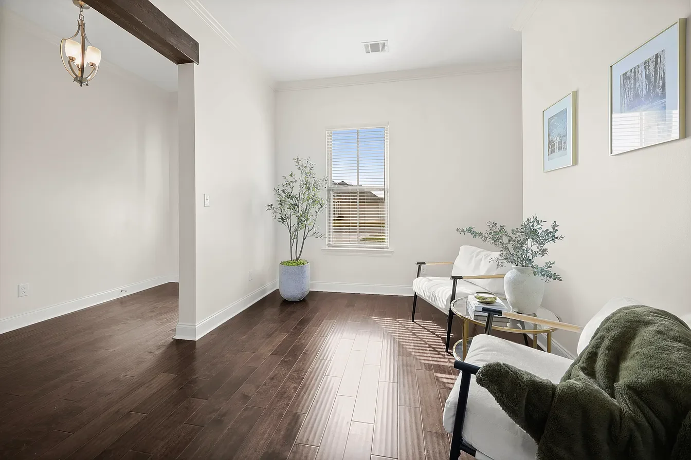 Bright living room with white walls, dark wood floor, a window with blinds, and minimal decor. Two white armchairs and a black armchair with a green blanket. A small round side table with a white vase and books. Two framed pictures hanging on the wall and a potted plant in the corner. Sunlight streaming through the window.