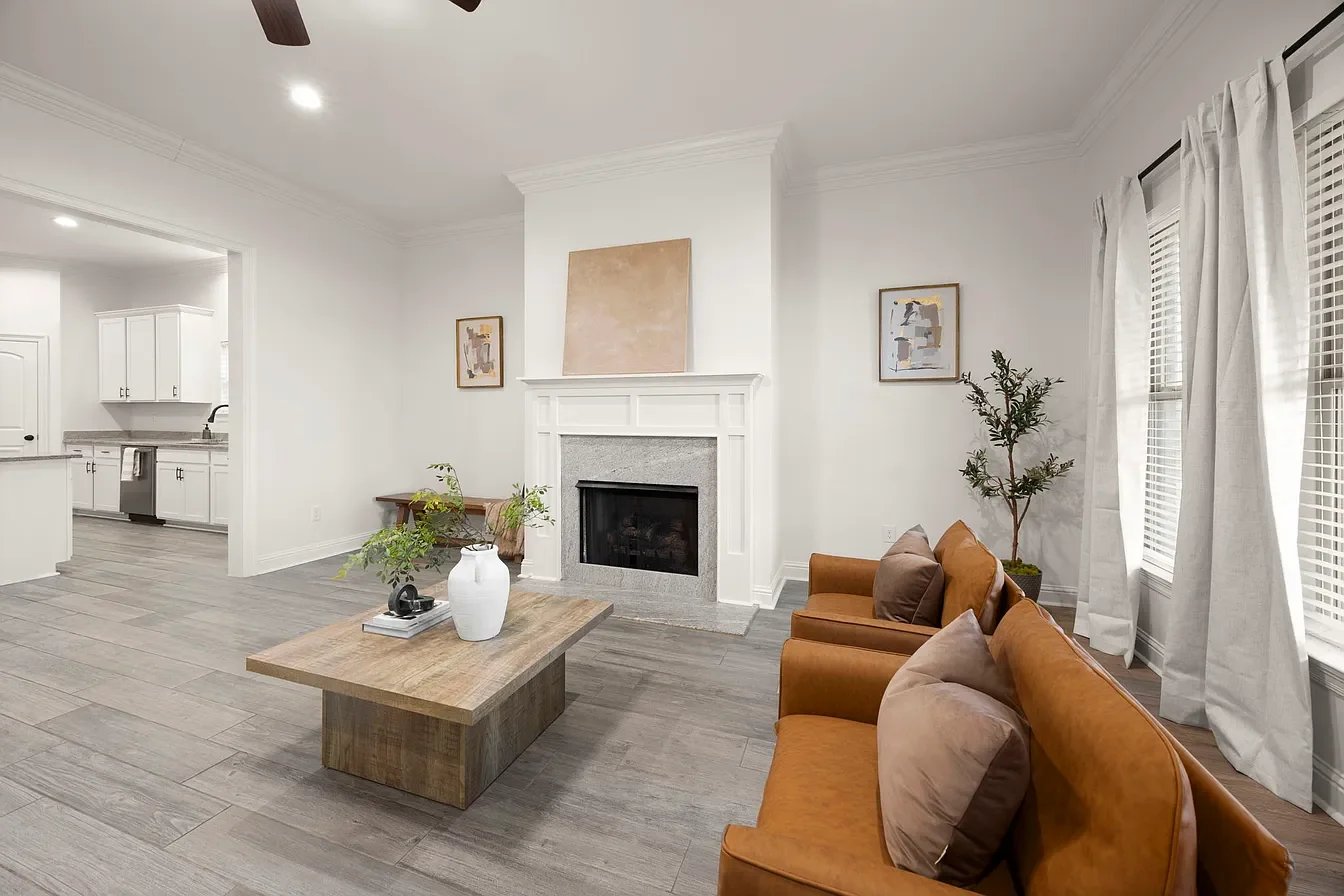A living room with a white fireplace, brown sofa with beige pillows, a coffee table with a white vase and green plants, white curtains, and framed artwork on the walls.