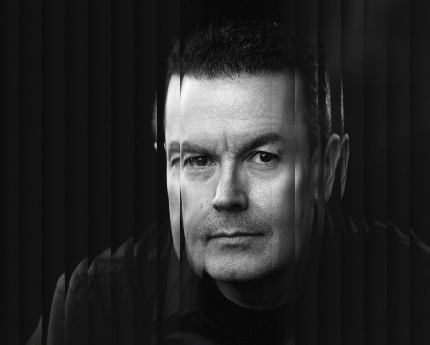 Black and white portrait of a man with short dark hair, looking directly at the camera with intense expression, through vertical black blinds.