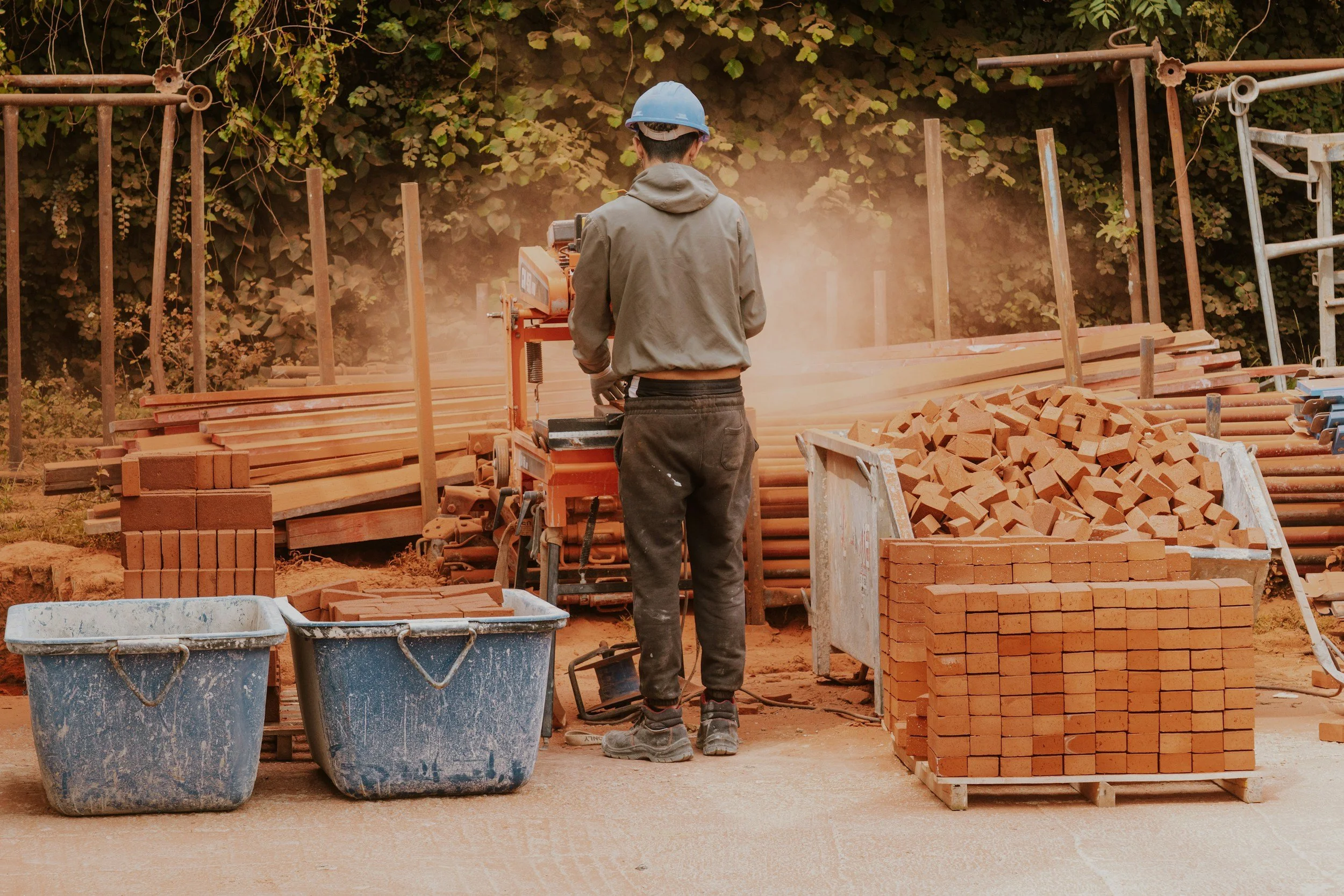 A construction worker using a saw to cut wood on a construction site with stacks of bricks, wood, and dust in the surroundings.