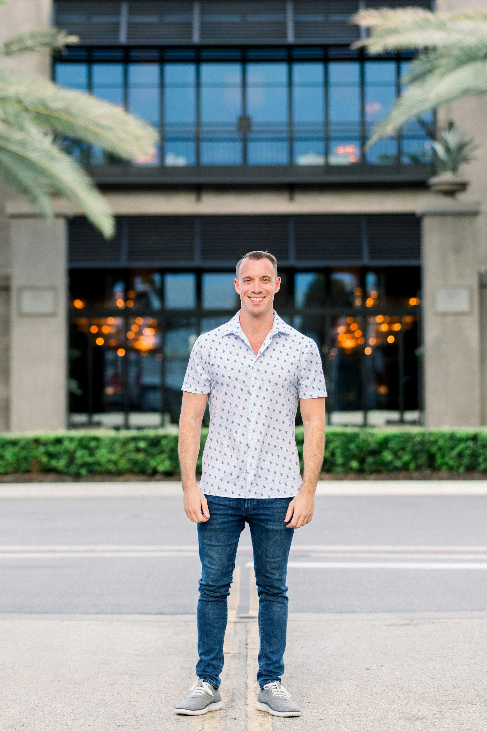 A smiling man standing in the center of the street in front of a modern building with large glass windows, palm trees on either side, and interior lighting visible through the windows.