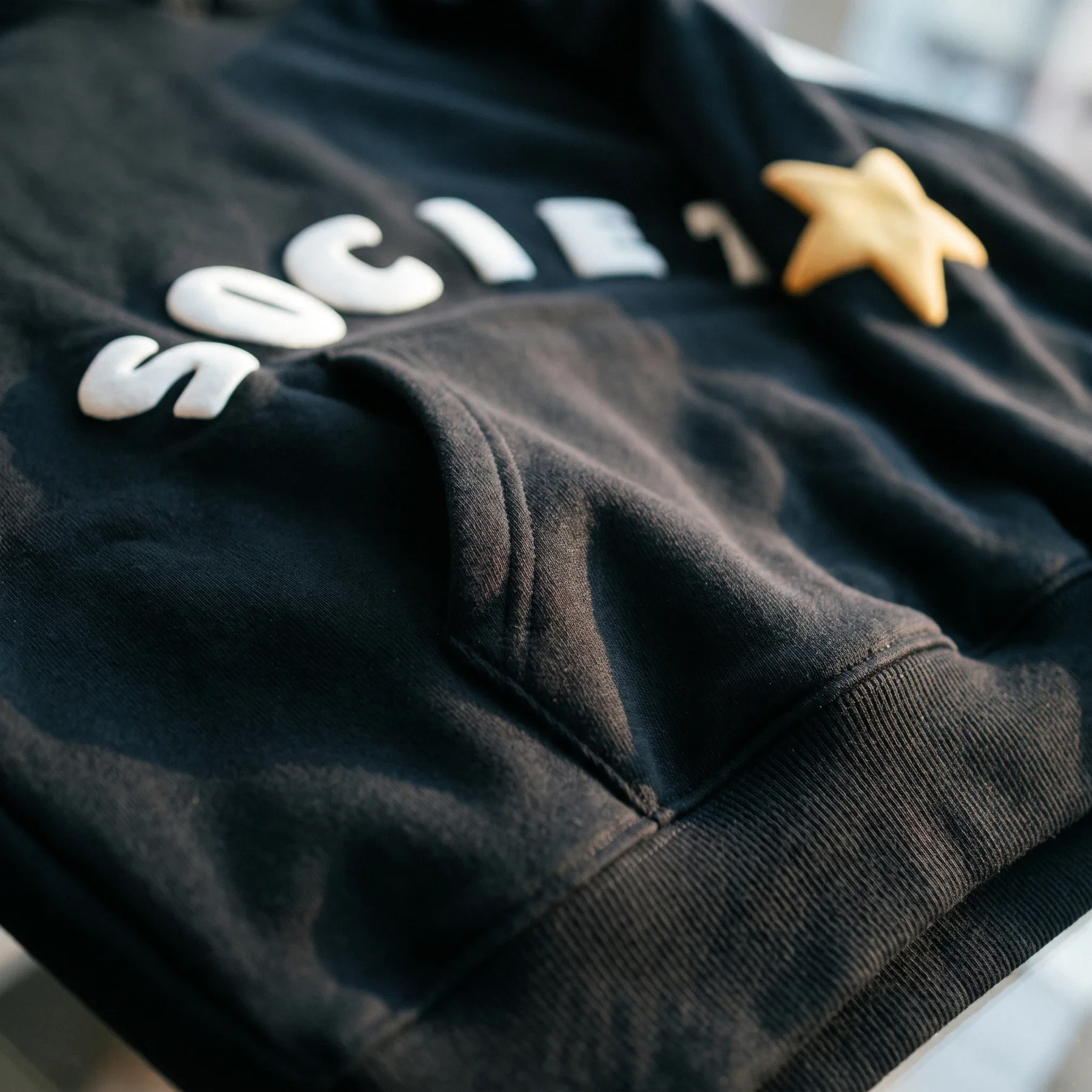 White puff SOCIETY lettering above Black Hoodie cotton, with ribbed hem and sleeve detail.