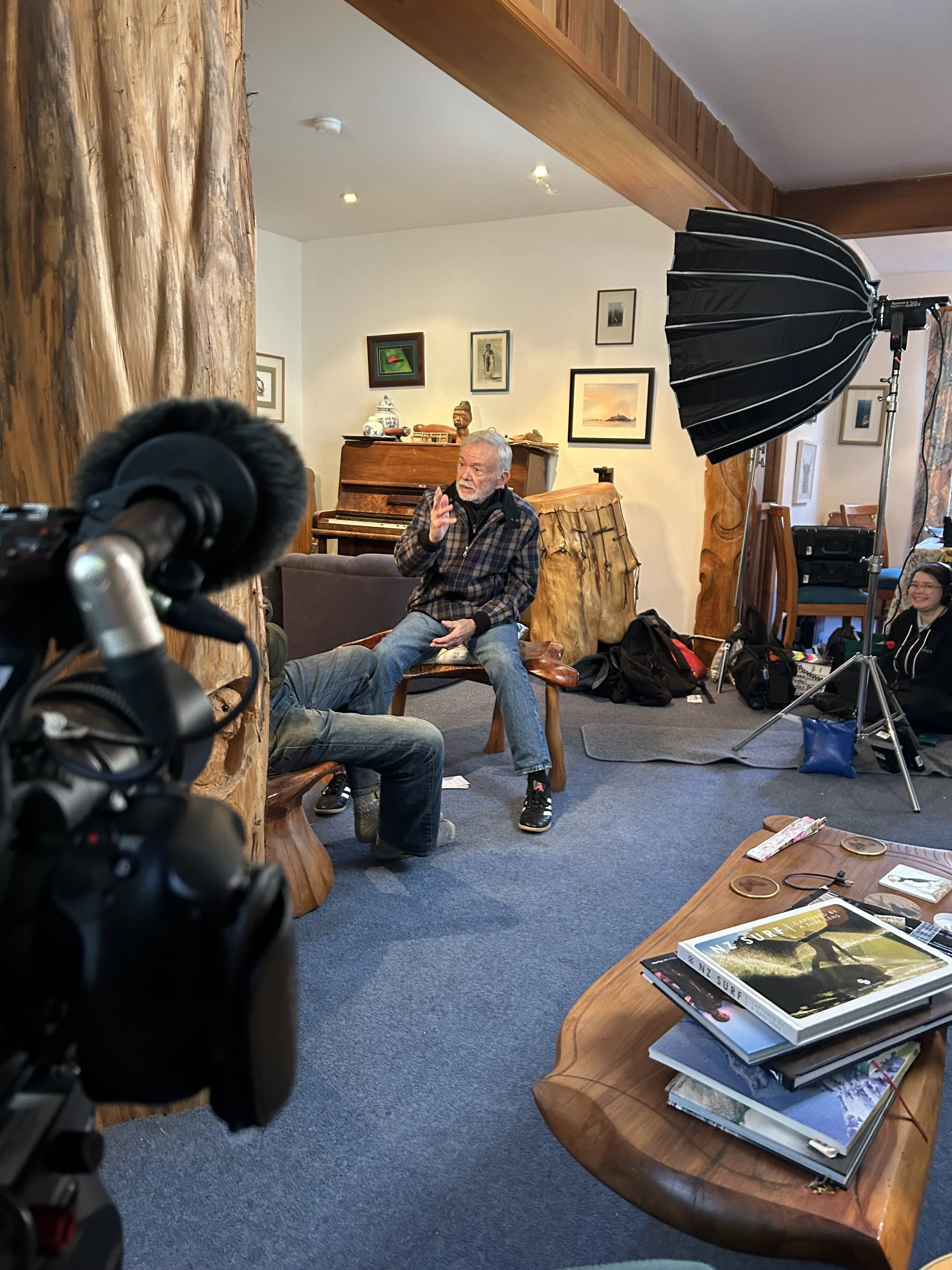 People filming a video interview in a cozy, wood-accented room with artwork on the walls, a wooden table with magazines, and production equipment including a camera and lighting.