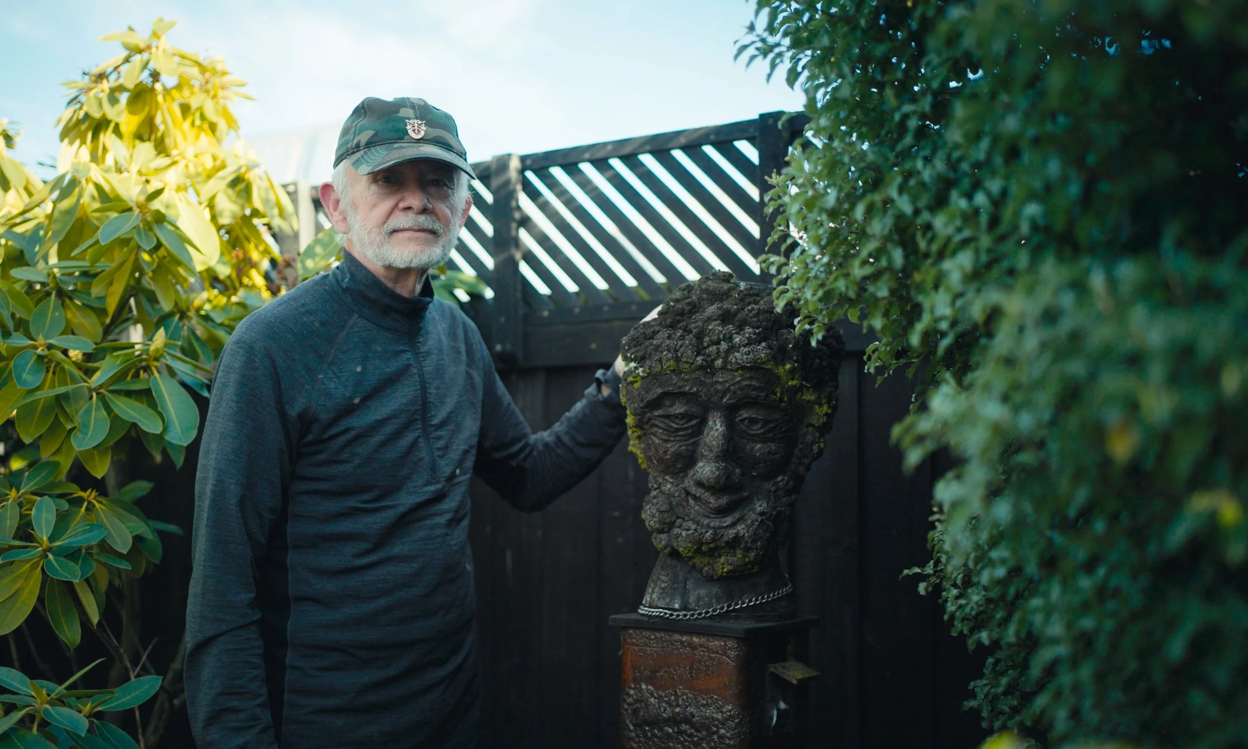 An older man with a white beard, wearing a gray cap and dark gray jacket, standing outdoors next to a large textured sculpture of a face with moss growth, in a garden with green foliage and a black fence in the background.