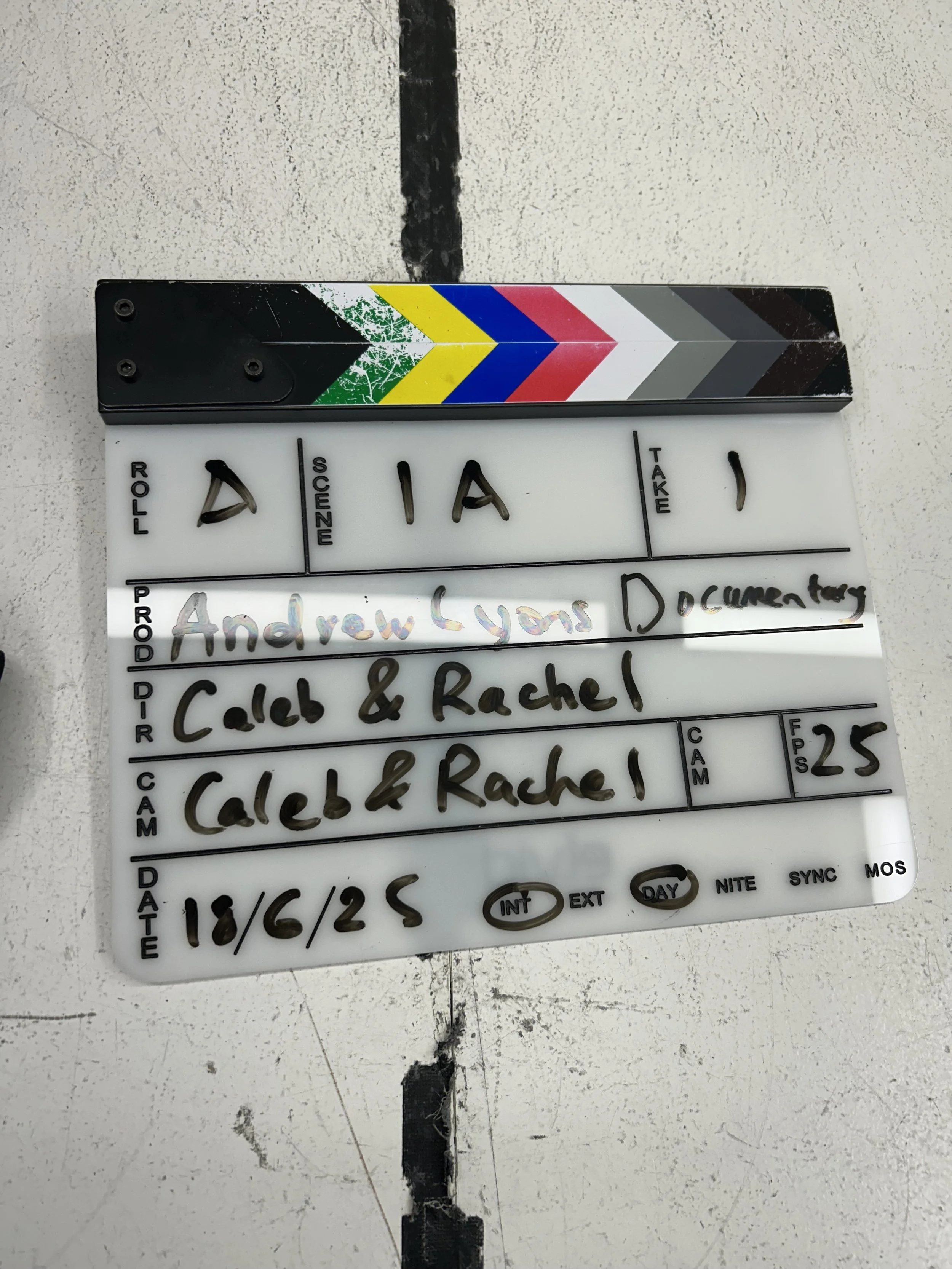 A film clapboard with handwritten details about a scene including the scene, the product or production title 'Andrew Lyons Documentary,' the names Caleb and Rachel.