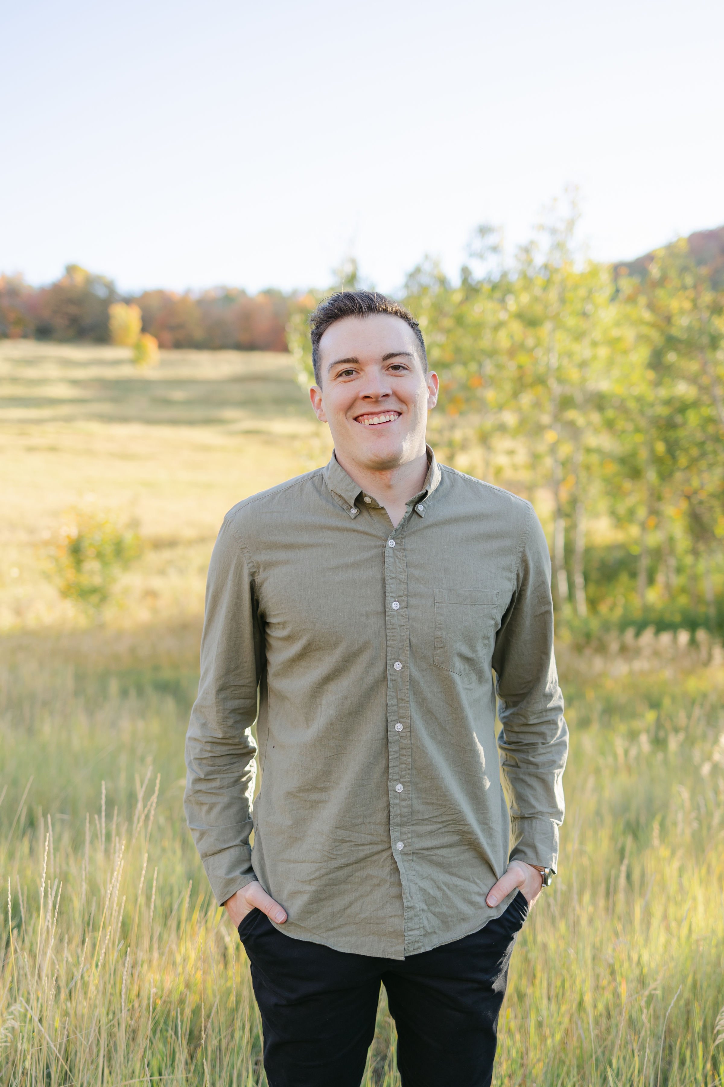 A young man standing in a field of tall grass with trees in the background during late afternoon or early evening.