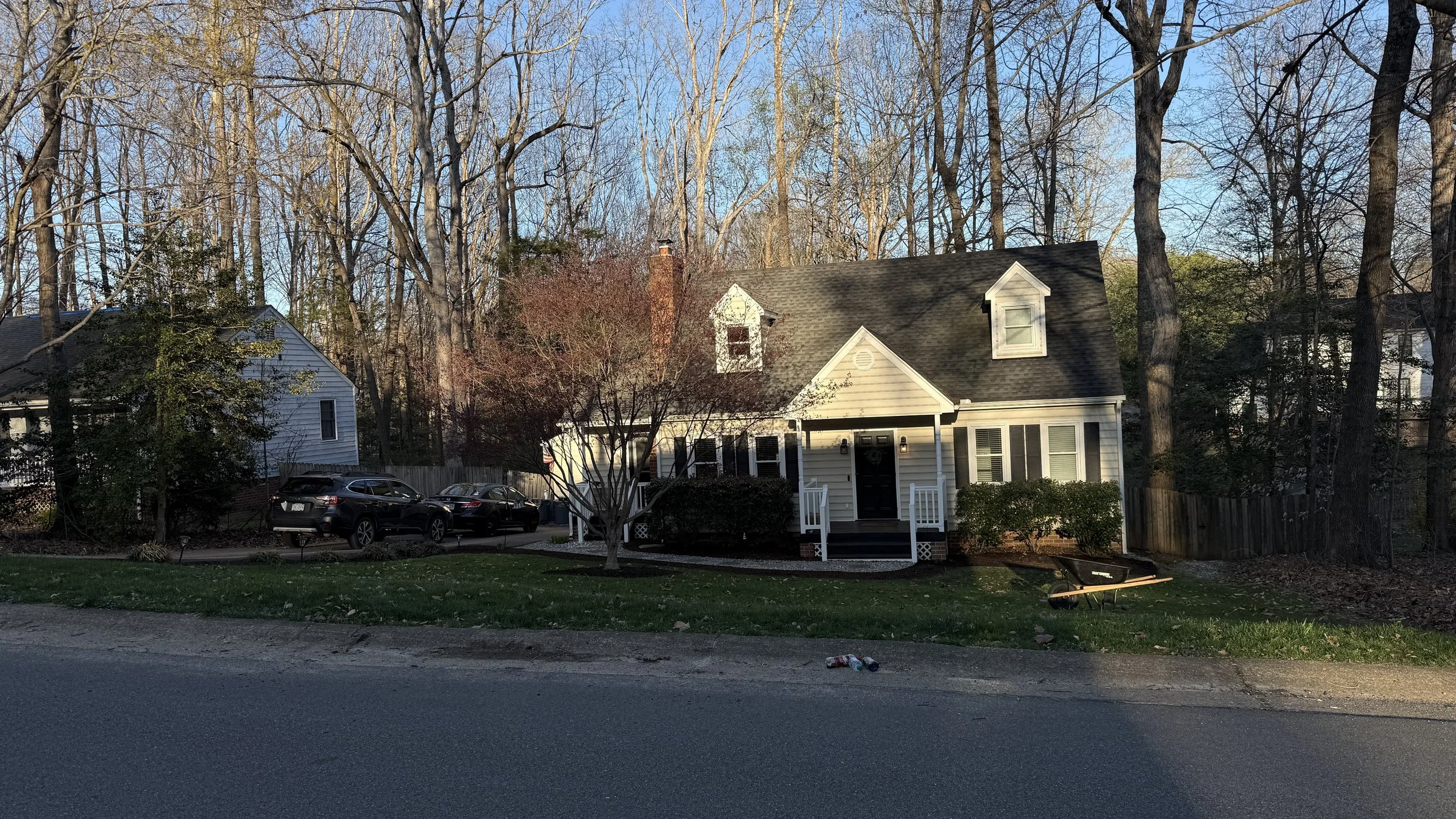 A white house with a black front door, small porch, and dormer windows surrounded by trees and parked cars in the driveway.