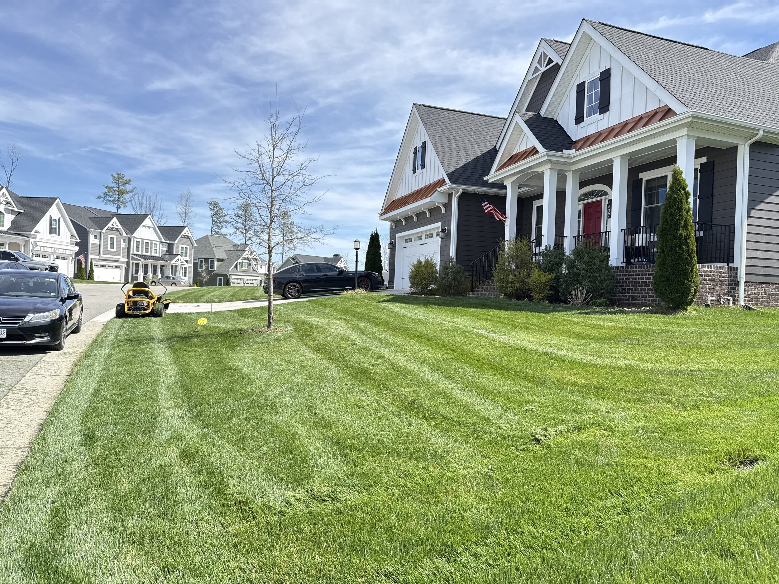 Upscale residential neighborhood with several modern houses, a paved street, parked cars, a lawnmower, a small tree with no leaves, and a well-maintained green lawn under a partly cloudy sky.