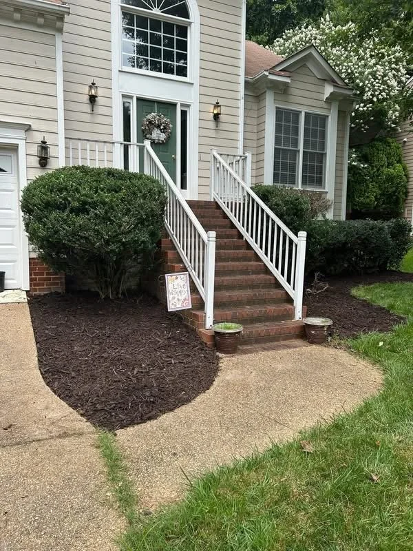 Front entrance of a suburban house with brick stairs and white railing, decorated with a wreath on the door, two wall-mounted lanterns, and a small sign on the ground. The yard is landscaped with mulch and bushes, with a concrete walkway leading to t