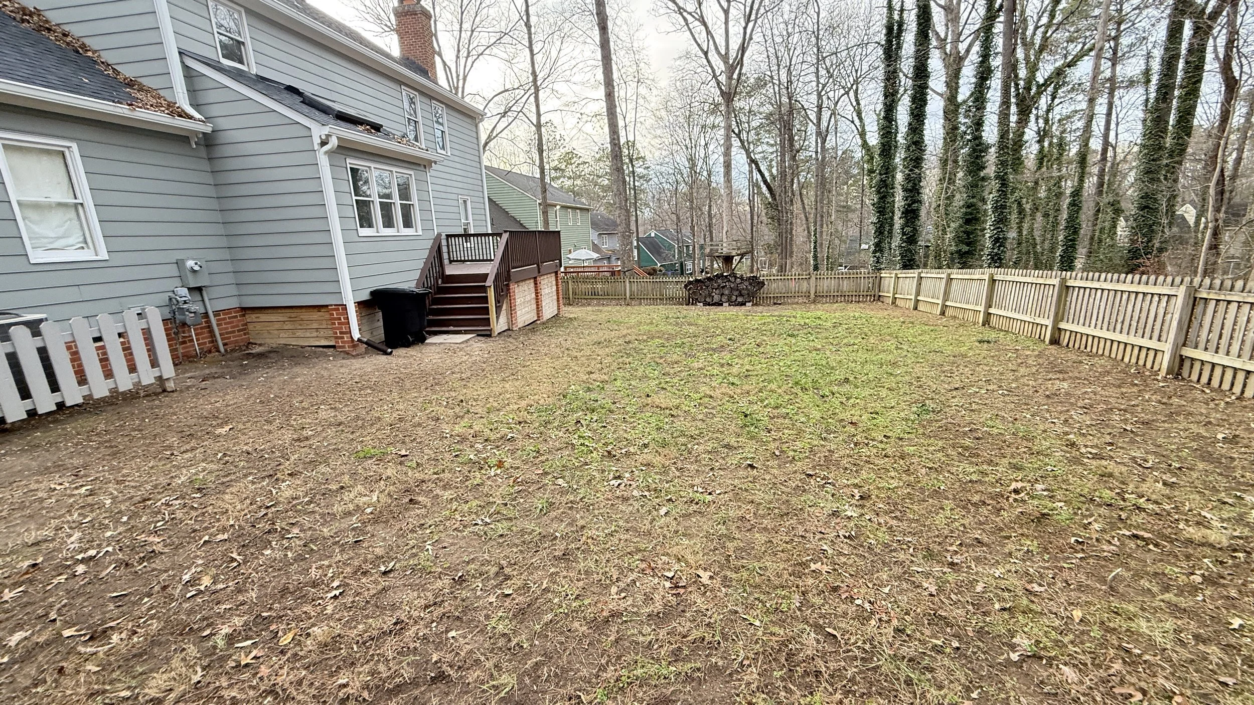 Backyard with a partially cleared lawn, wooden fence, and trees in the background. The house has gray siding, white windows, and a small deck with stairs.