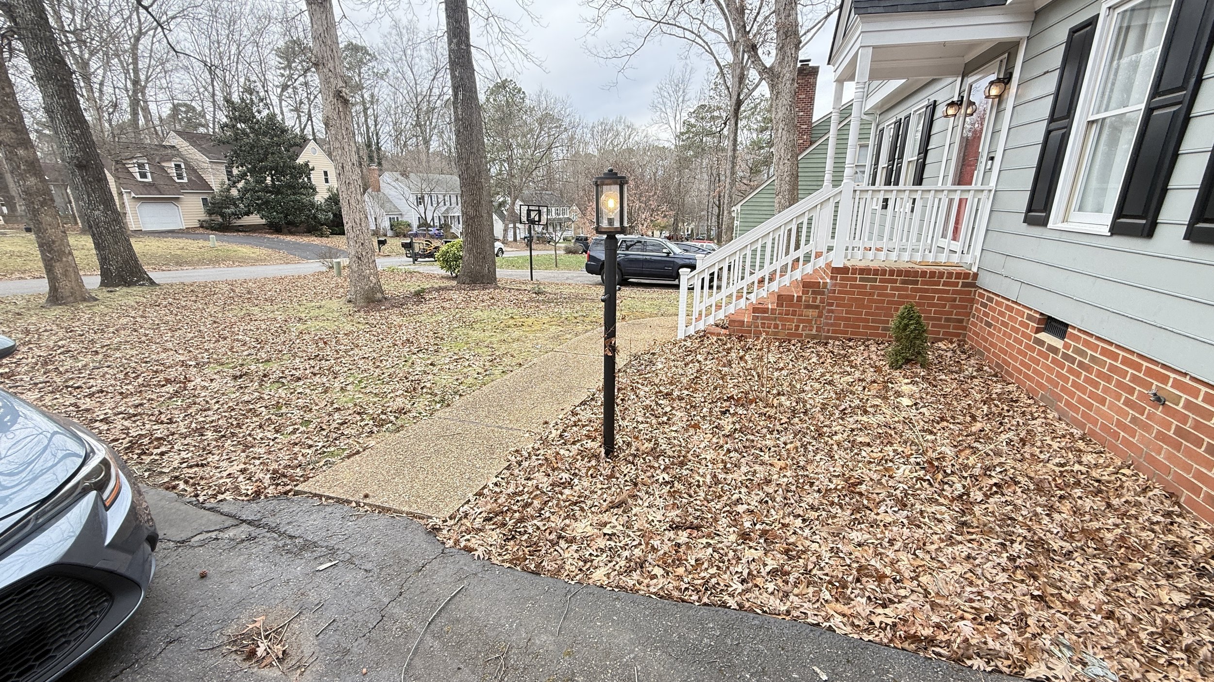 The front yard of a house with fallen leaves, a concrete walkway, a street lamp, and neighboring houses in the background.