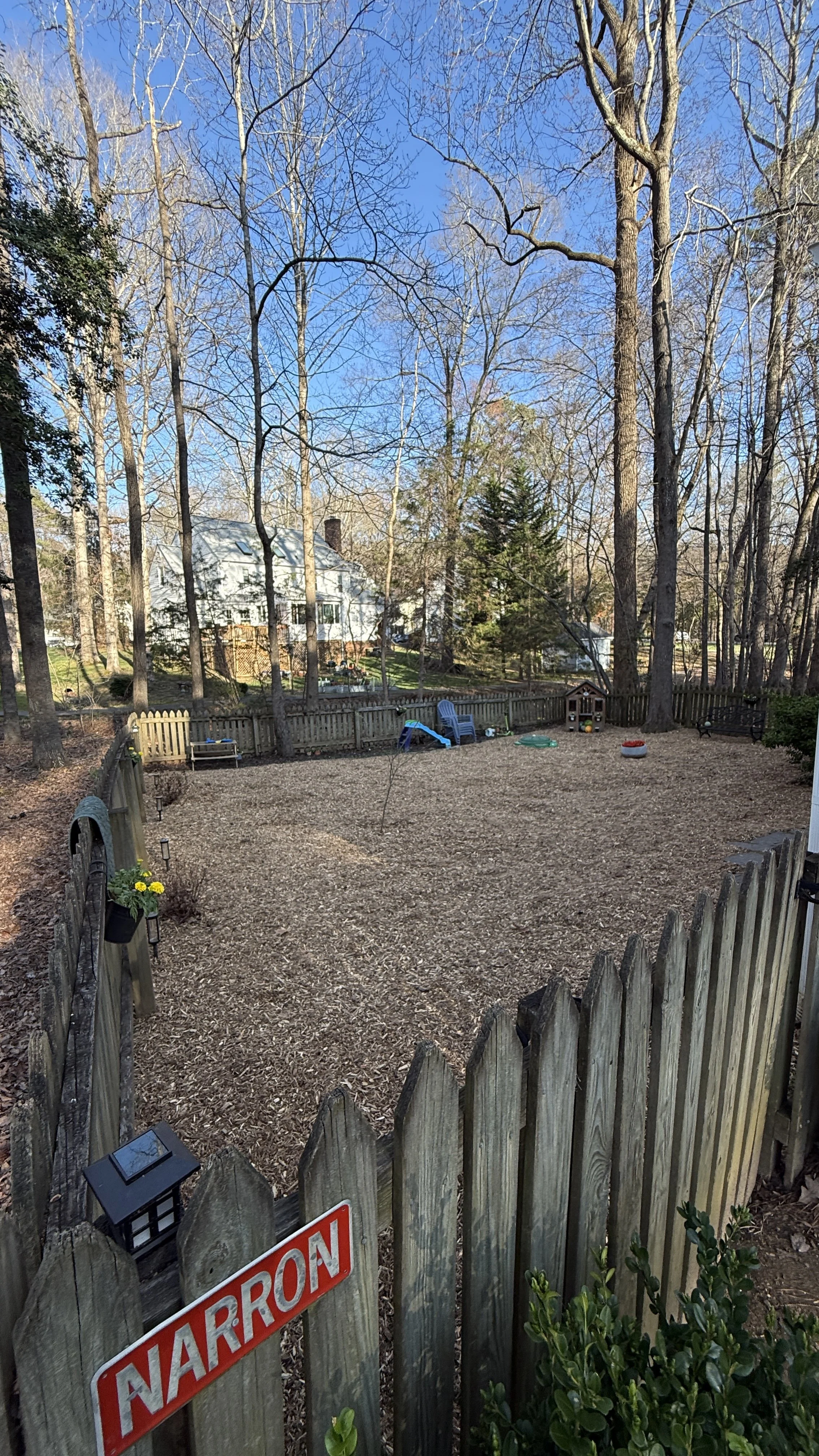 A fenced backyard with a wood chip ground cover, surrounded by tall leafless trees and a wooden picket fence. There are outdoor chairs, a small playhouse, and toys scattered around. A house with a chimney is visible in the background through the tree