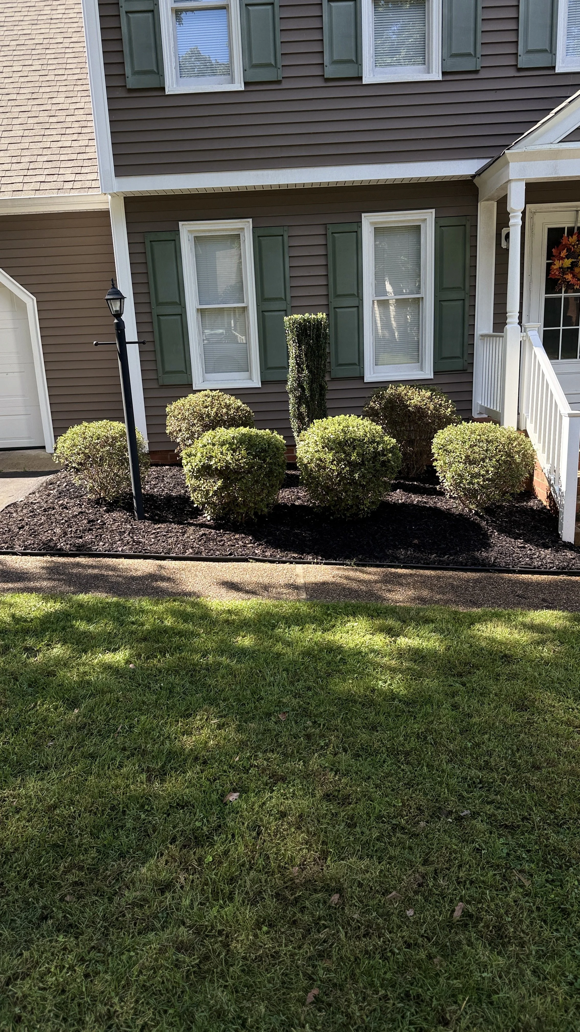 Front yard with green bushes, mulch bed, sidewalk, and a house with brown siding, green shutters, white windows, and a porch.