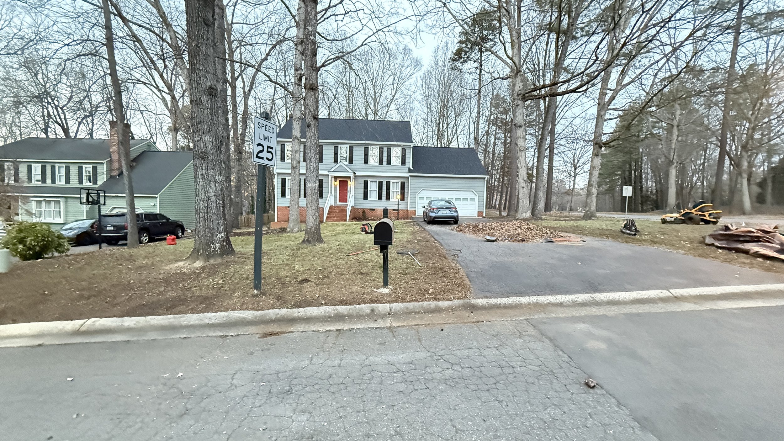 A two-story house with a white exterior, black shutters, and a red front door. The house has a driveway with a gray car parked on it. There are tall trees in the front yard and a speed limit sign showing 25 mph. There is a mailbox in the yard and som