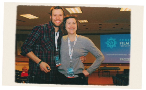 Dan and Rachel standing together smiling in an auditorium. A large screen in the background reads "Frozen River Film Festival". They are also wearing lanyards with Frozen River Film Festival badges around their necks.