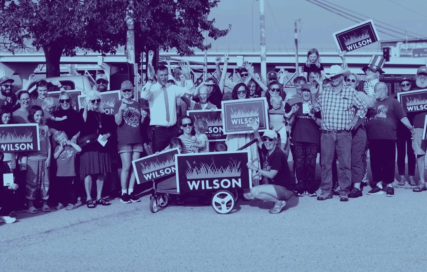 Dan lifting his hands up and smiling. Winona County residents are around him holding election signs and waving.