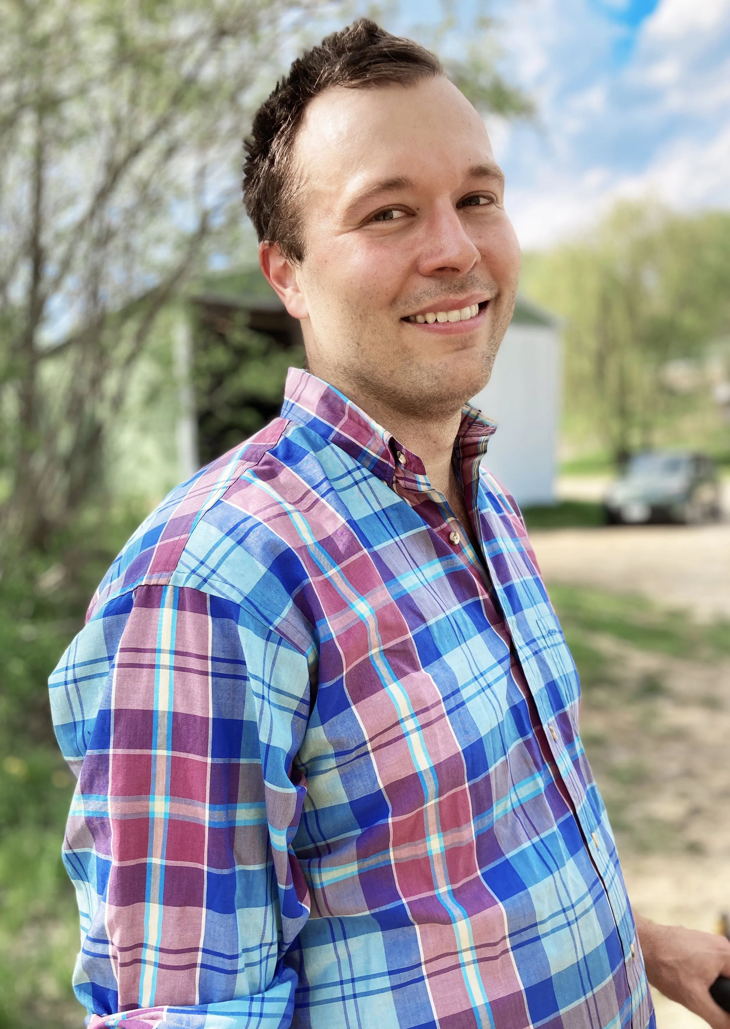 Dan Wilson smiling and looking into the camera. He's wearing a plaid blue and purple shirt. He's standing outdoors in Minnesota with blue sky and green trees behind him.