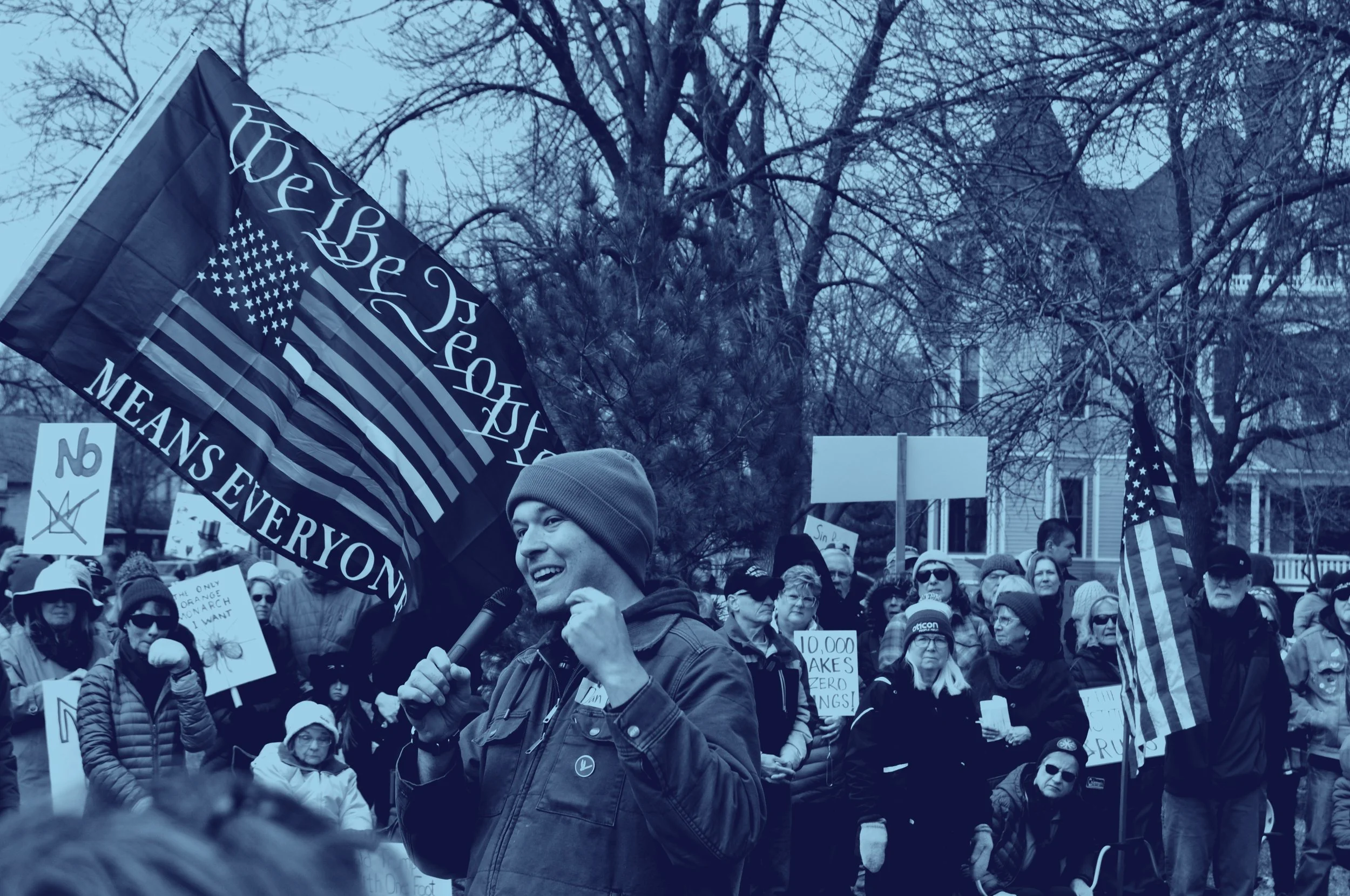 A picture of Dan Wilson giving a speech at the No Kings rally with Winona residents around him. A flag flies above with a rainbow USA flag and it reads "We The People Means Everyone".