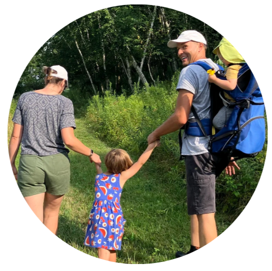 Dan and Rachel walking on a forest trail holding hands with one of their children. The other is riding on Dan's back. Everyone is facing away from the camera. Dan is looking over his shoulder, smiling.