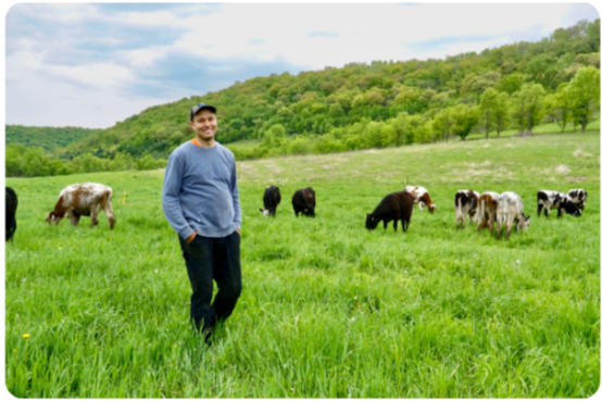 Dan Wilson wearing jeans, long sleeve shirt, and a baseball cap. He's smiling in a green pasture with his cows behind him.