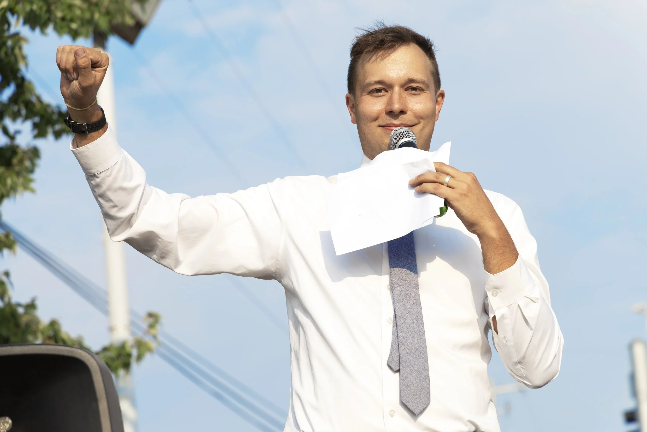 Dan holding a raised fist in the air during his speech. He's smiling into the camera. A blue sky is behind him.