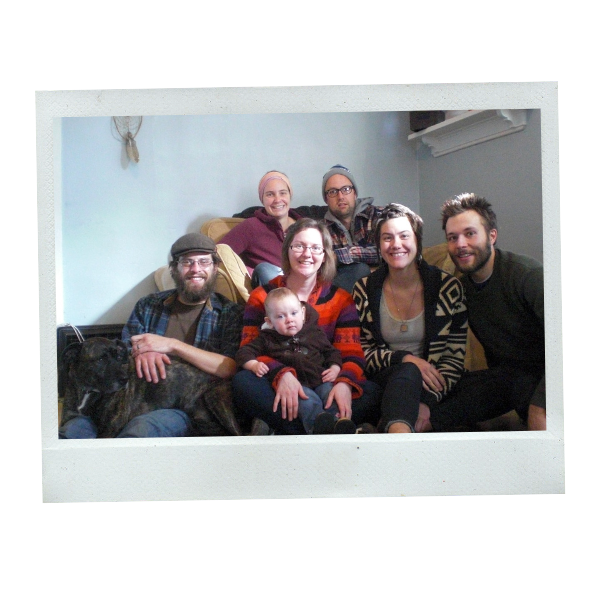 A group of happy friends sitting indoors. They're on a couch, smiling at the camera. One man has a dog in his lap. The woman in the middle has a baby in her lap. Rachel and Dan are posed together with them.