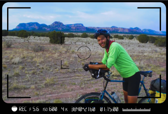 Dan Wilson wearing a bright green shirt, biking shorts, and a helmet. He's on a bike with an Arizona landscape behind him.
