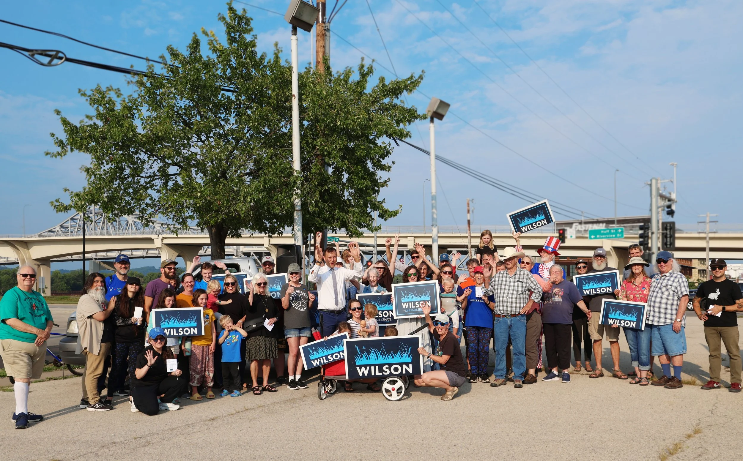 Dan Wilson during his Minnesota State House election announcement. He's outdoors, wearing dark blue pants, a white collared shirt, and a light blue tie. He's surrounded by members of Winona County holding Wilson for MN State House signs.