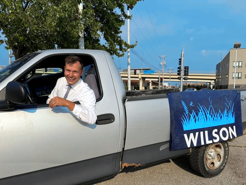 Dan parked in a gray truck with a Dan Wilson blue banner on the back. His head is out of a rolled down window and he's smiling at the camera.