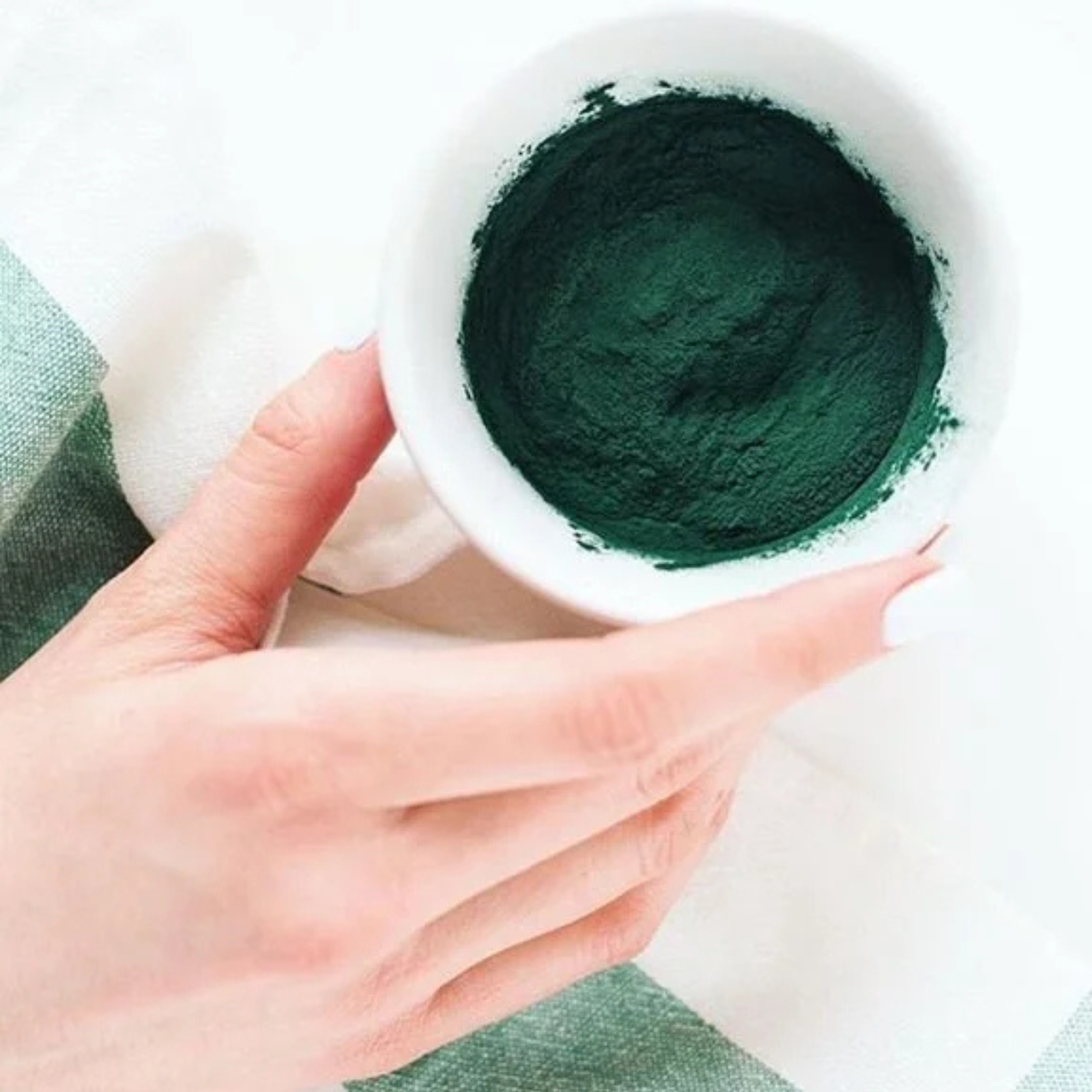 A person's left hand holding a small white cup filled with dark green powder, with the person's thumb and index finger visible. The background includes a white surface and a green fabric.