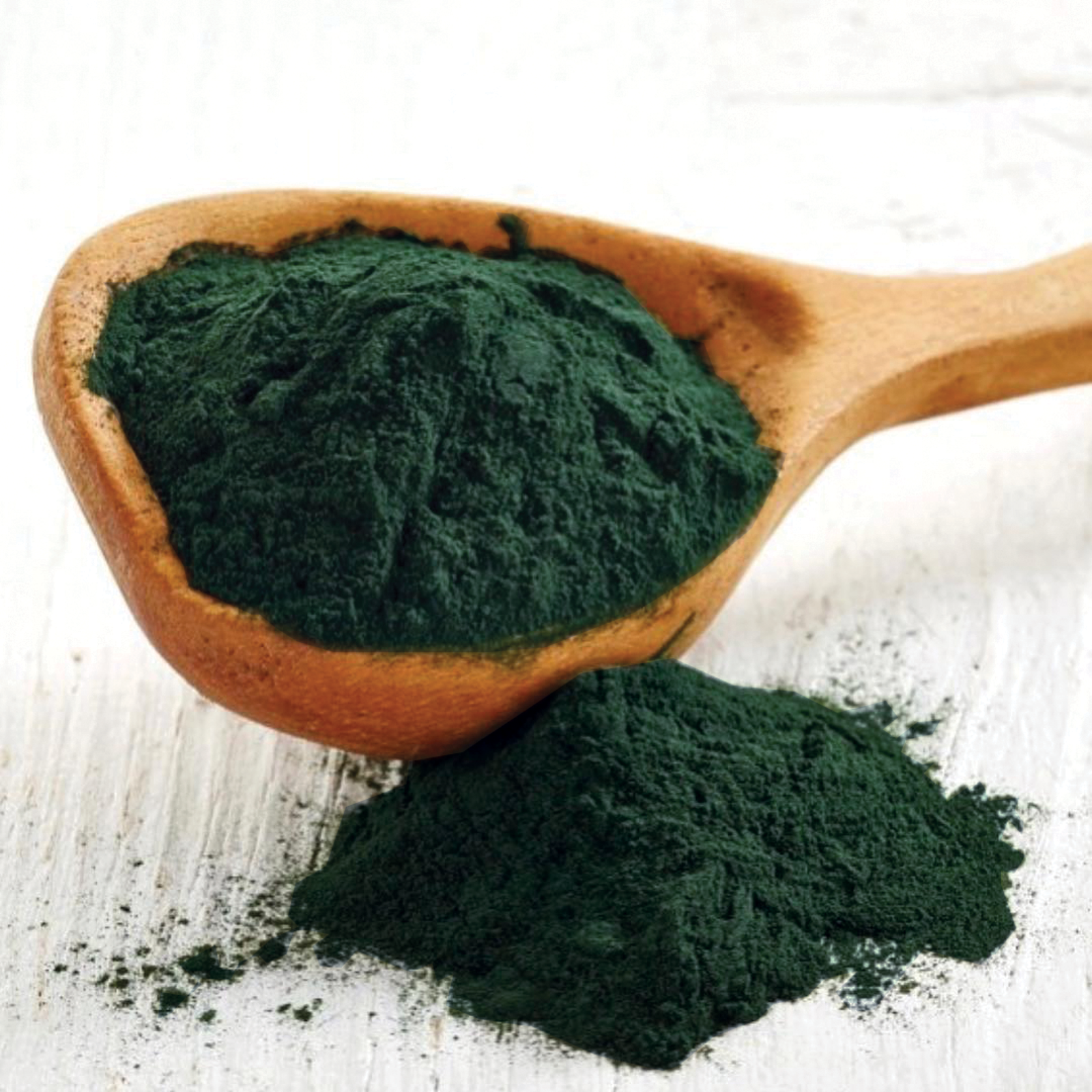 A wooden spoon filled with dark green powdered substance and some powder spilled on a white surface.