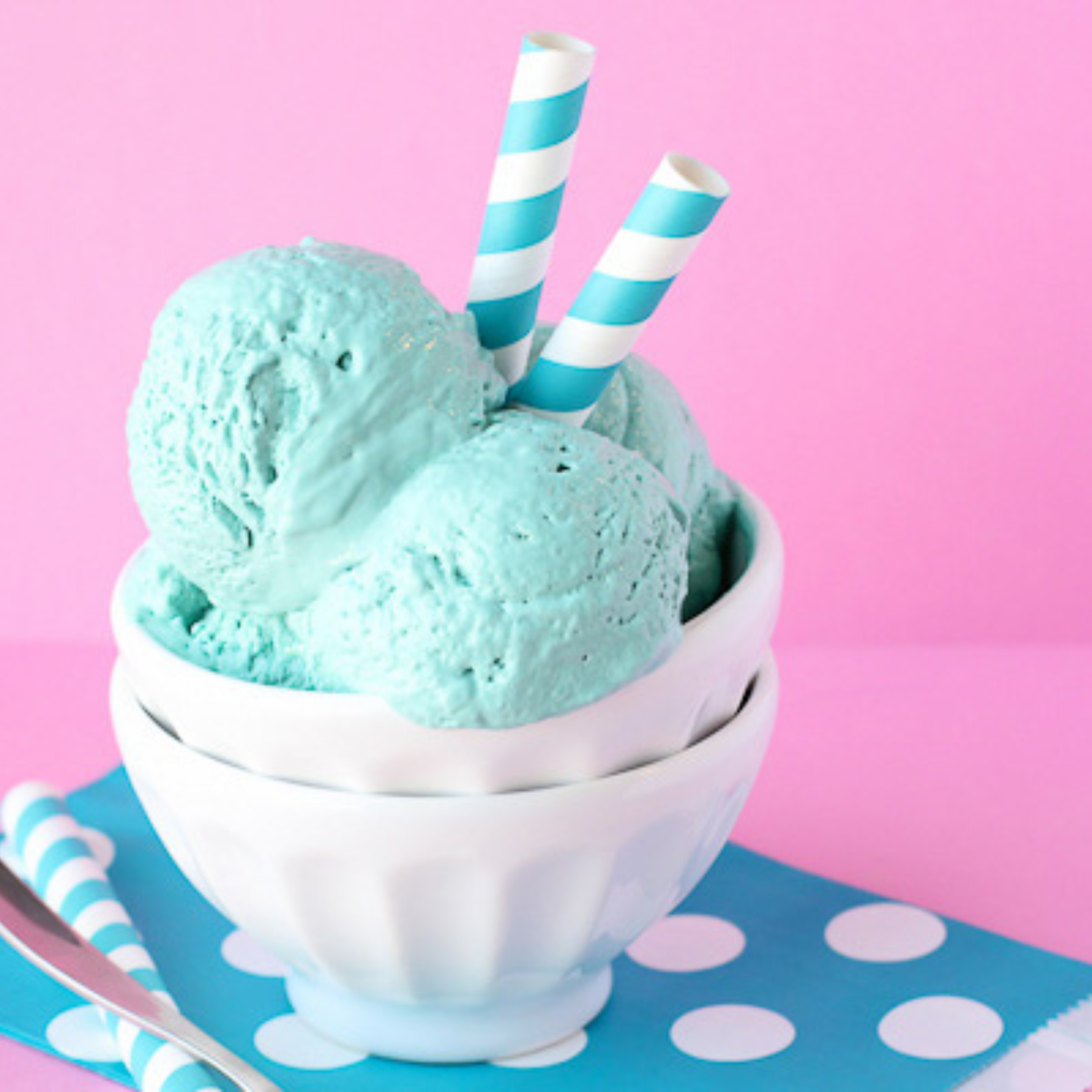 A bowl of mint ice cream scoops with two blue and white striped paper straws, set on a blue and white polka dot napkin against a pink background.