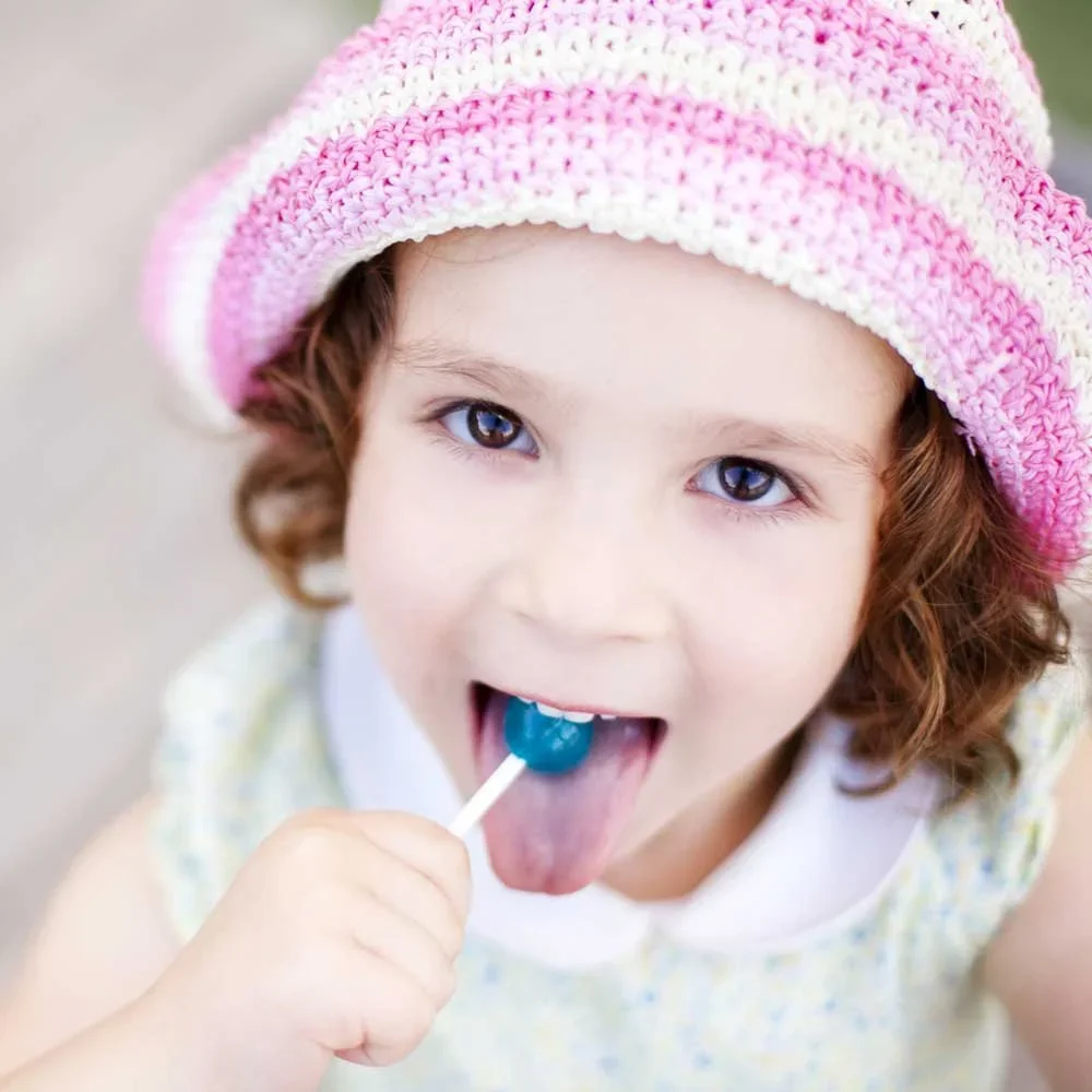 A young girl with curly brown hair wearing a pink and white striped knitted hat, licking a blue lollipop.