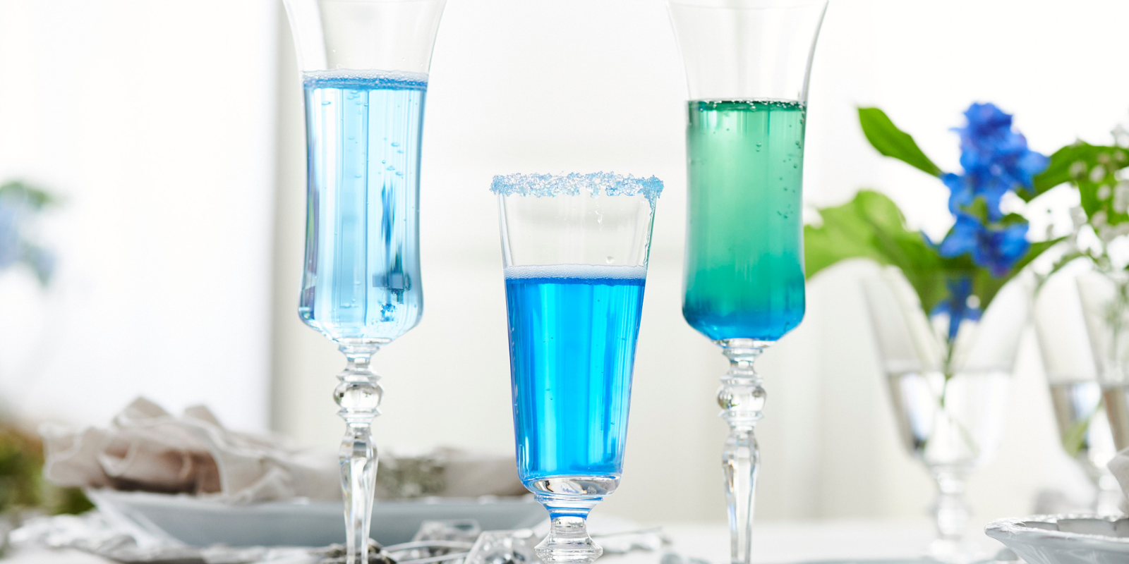Three champagne flutes with colorful blue and green drinks, and a vase with blue flowers in the background.