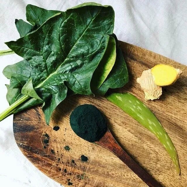 Fresh spinach leaves, a green aloe vera leaf, a piece of ginger with yellow interior, and a mound of black powder on a wooden cutting board.
