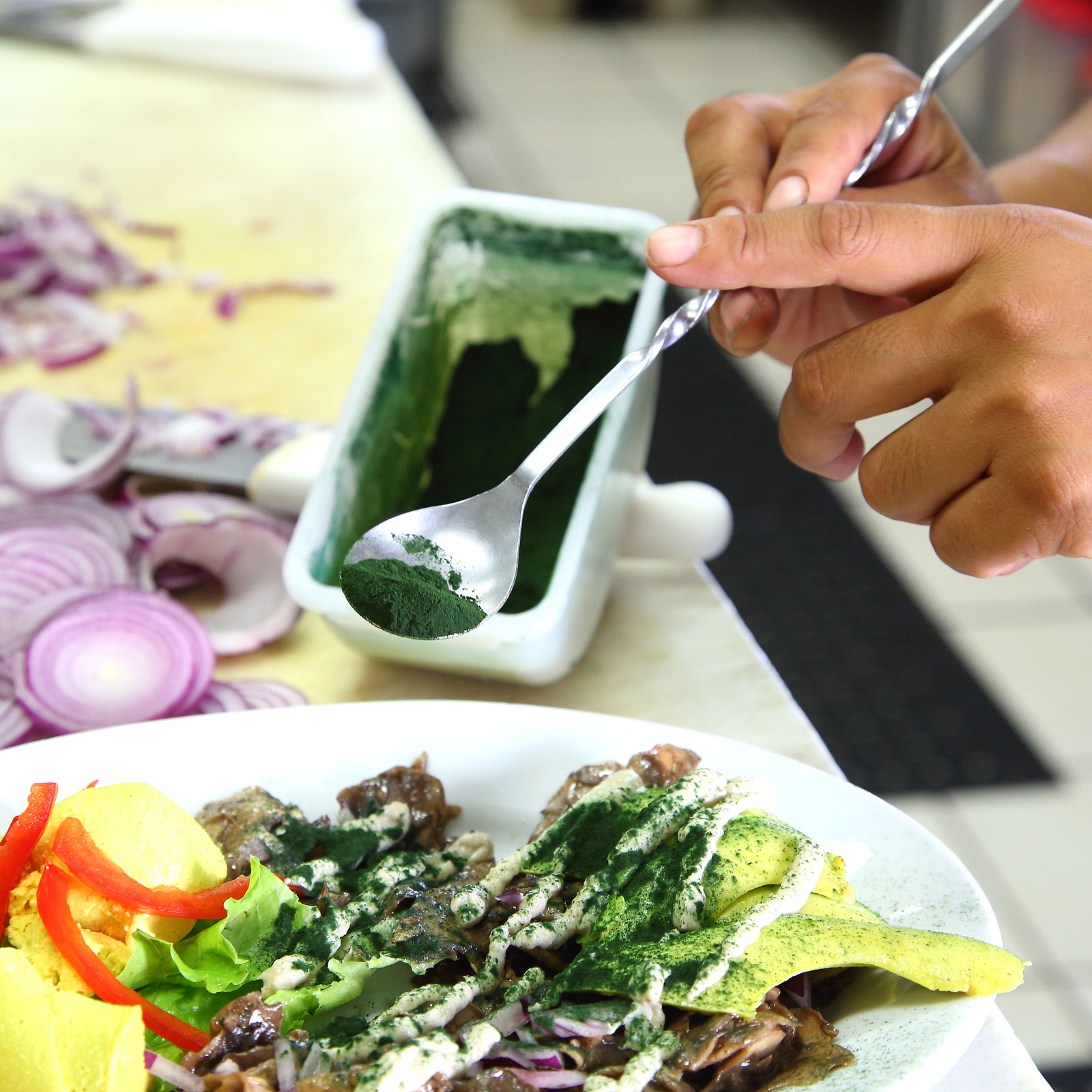 A person adds green powder to a dish of salad with sliced red onions, pepper, and lemon slices. The salad is garnished with a creamy dressing and vegetables.