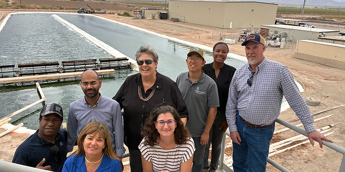 Group of nine people standing and sitting by a large water treatment pond in an industrial or research facility, with buildings and equipment in the background.