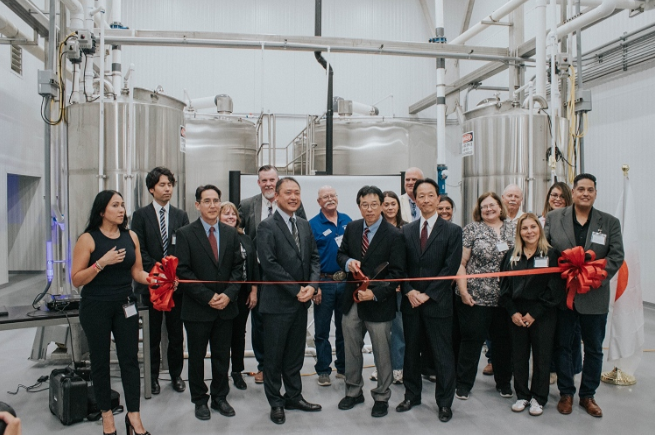 Group of people at ribbon-cutting ceremony in industrial setting with large stainless steel tanks in background.