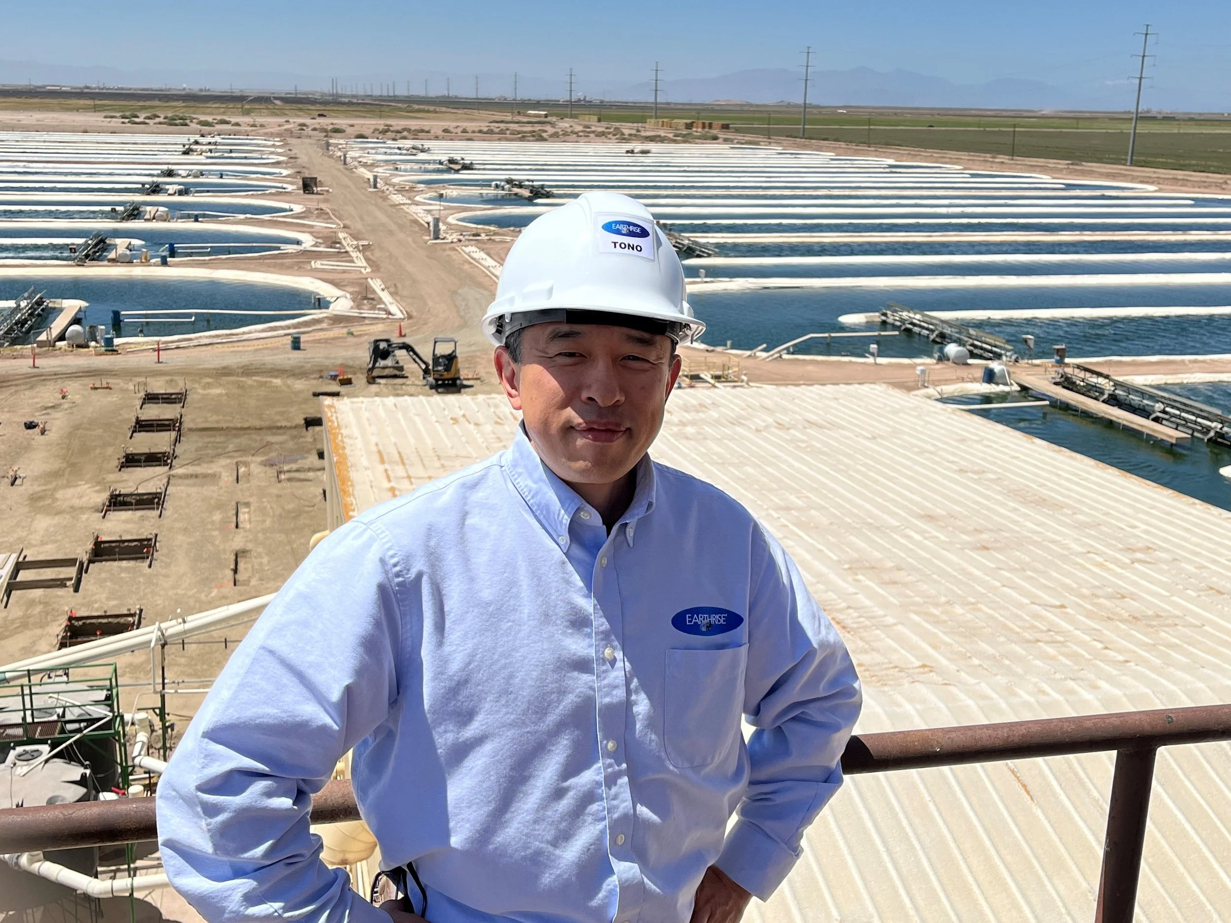 Man wearing a white hard hat and a light blue shirt standing on a balcony overlooking a large water treatment facility with multiple circular and rectangular tanks in the background.