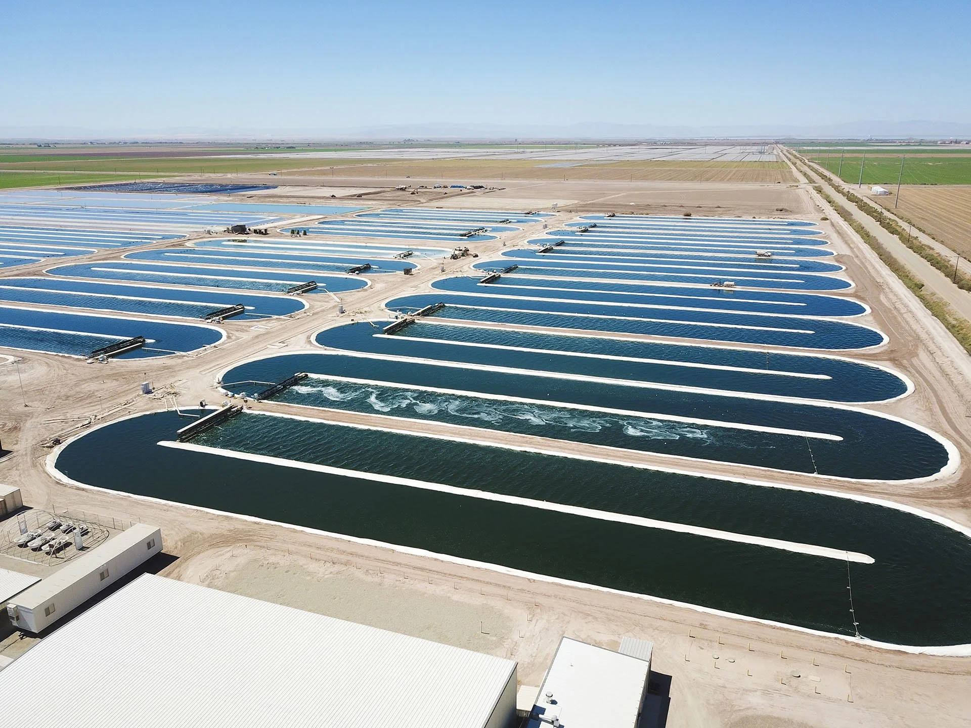 Aerial view of a large solar panel farm with water ponds for solar thermal energy. The water ponds are oval-shaped and lined with white borders, positioned in rows across a desert landscape. Several small structures and equipment are visible around the ponds.