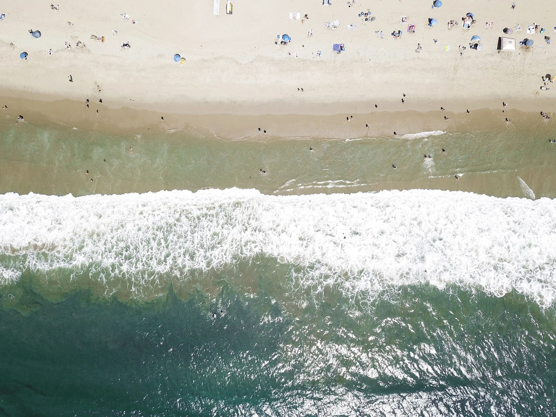 Aerial view of a beach with people swimming and sunbathing on the sandy shore, umbrellas, and beach chairs, with waves crashing onto the sand.