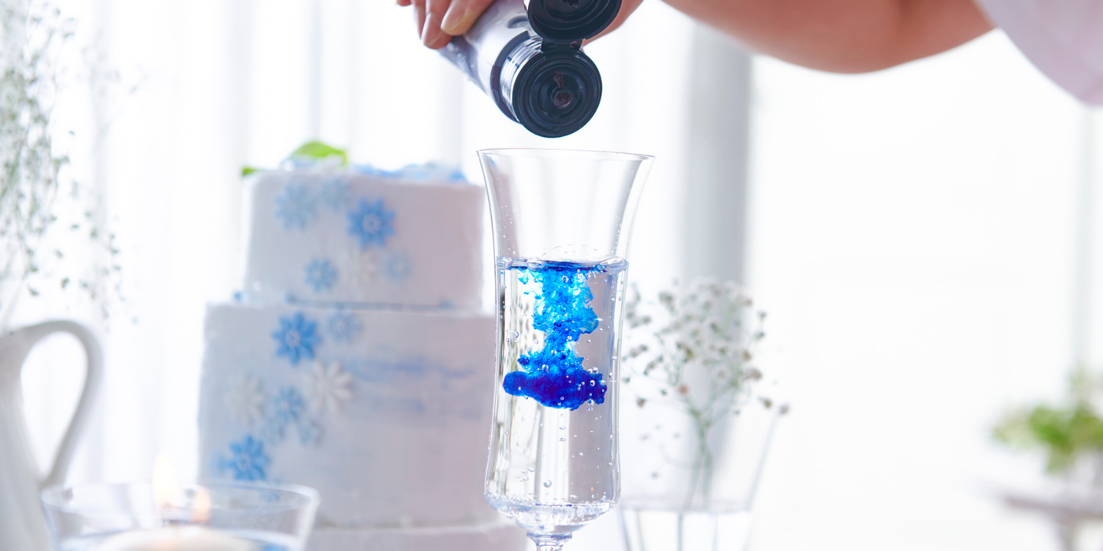 Person pouring blue dye into a tall glass of water with a wedding cake in background.