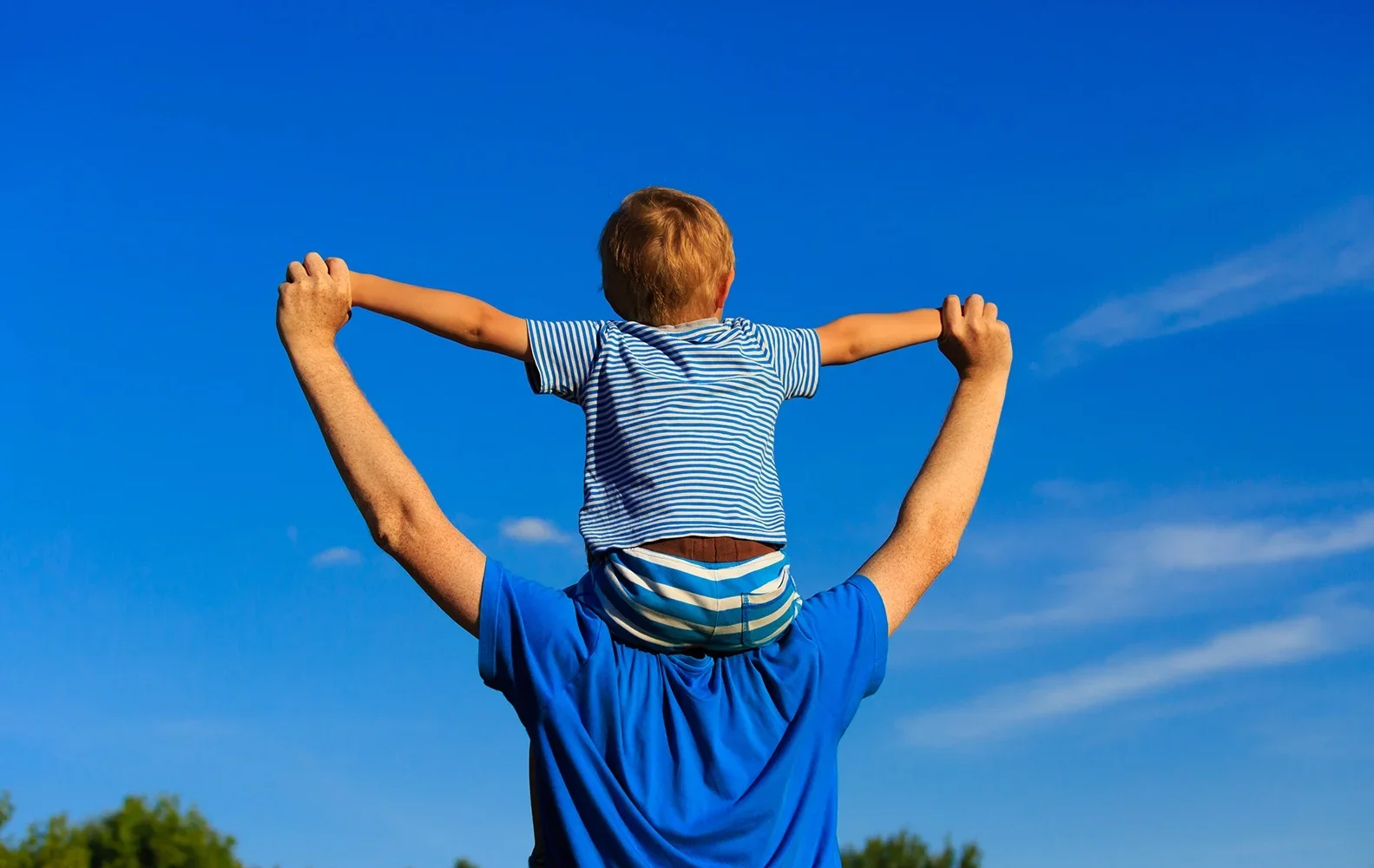 A man lifting a young boy on his shoulders outdoors against a bright blue sky, with the boy extending his arms outward.
