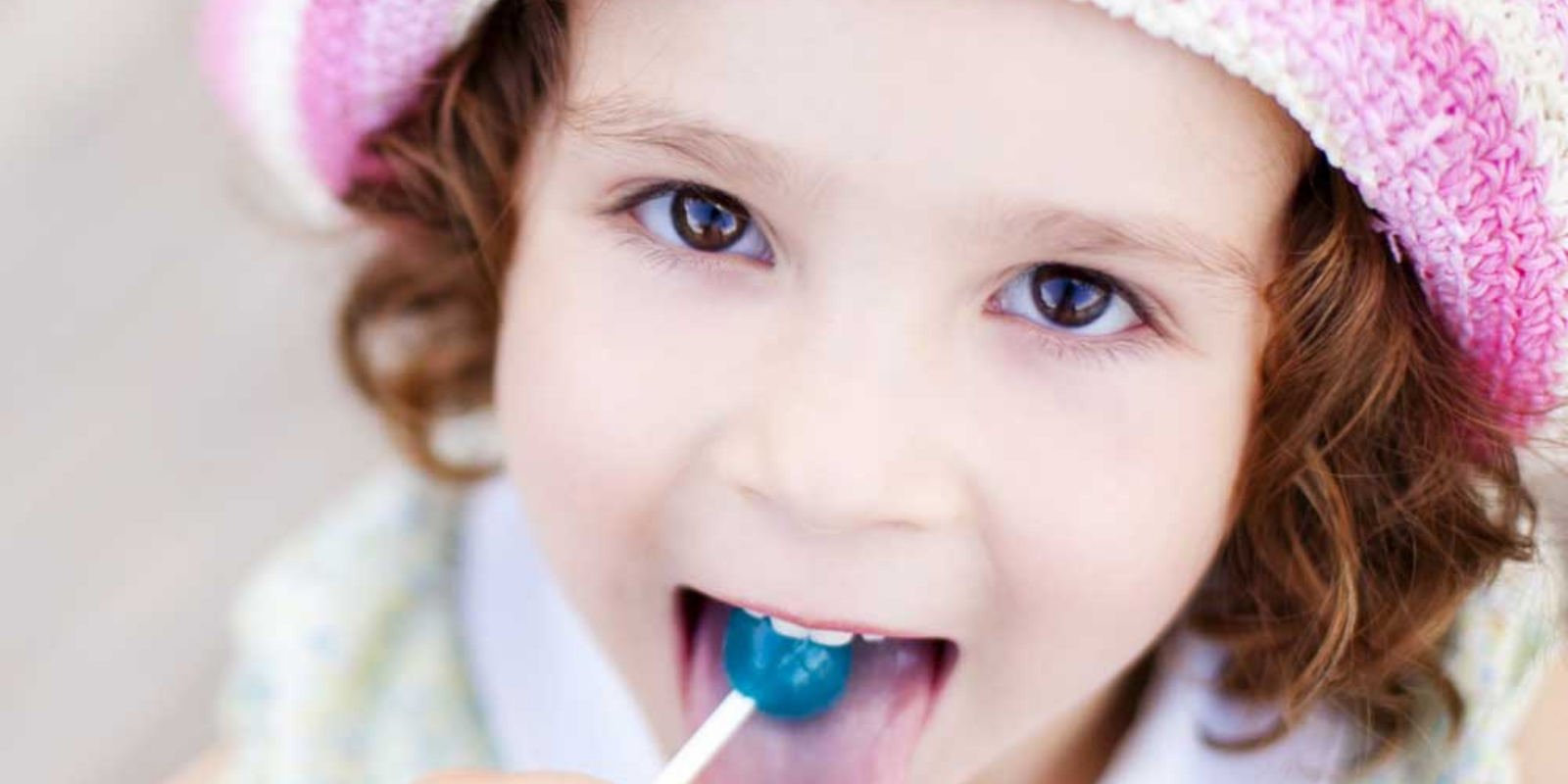 Close-up of a young girl with curly brown hair wearing a pink and white knit hat, licking a blue lollipop.