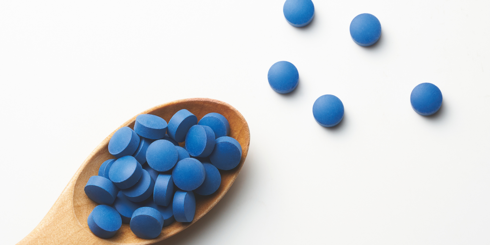Blue tablets or pills in a wooden spoon with some spilled onto a white surface.