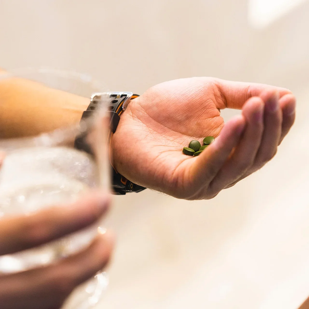 Hand holding three green pills, with a blurred glass held in the other hand in the foreground.
